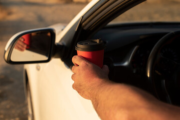 A hand holding a red coffee cup extends from a white car window. The scene captures a moment of...