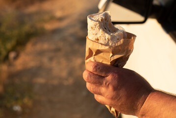 A hand holding a partially eaten fast food. The background is blurred, suggesting a warm outdoor...