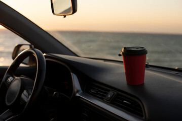 A red coffee cup sits on the dashboard of a car, with a view of the ocean at sunset. The scene...