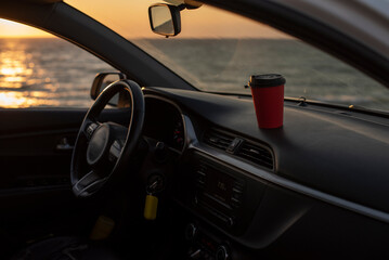 Interior of a car with a red coffee cup on the dashboard. The ocean is visible in the background...