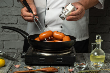 A chef prepares sausages in a black frying pan, adding salt and spices while standing at a rustic...