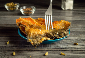 Fried fish is presented in a blue bowl, with a fork poised to serve a piece. A set of spices...