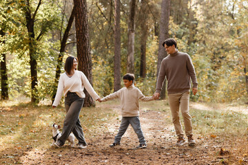 Front view of a smiling Caucasian family walking hand in hand with their small dog in an autumn...