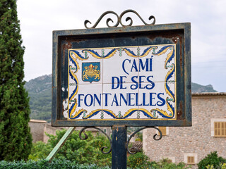 Obraz premium Decorative ceramic street sign for Cami de ses Fontanelles in Soller, Mallorca, framed in wrought iron with scenic mountain backdrop.