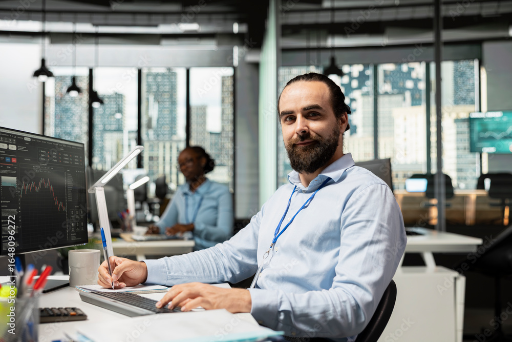 Poster portrait of smiling trader taking notes while analyzing candlestick charts and price graphs on scree