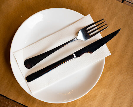 In refreshment bar on wooden table, paper tablecloth round plate with cutlery, knife and fork complemented by transparent glass for drinks. Restaurant serving option