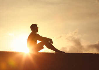 thoughtful man sitting on grass looking out to the beautiful sunrise with happy feelings, peace of...