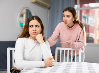 Upset young woman sitting at home table with disgruntled woman behind