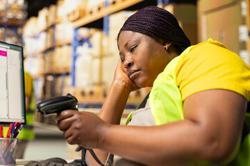 African american female napping on warehouse desk while she uses scanner, scanning shipping labels...