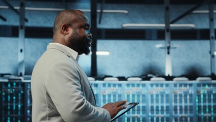 Technician working in high tech server hub, analyzing data on tablet to ensure seamless data flow. IT expert examining equipment in data center, ensuring system integrity and security, camera B