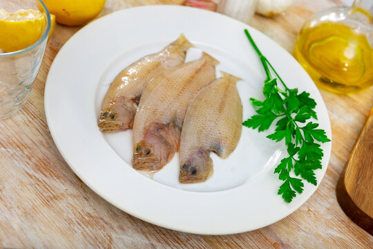 Whole raw scaldfish on white plate surrounded by fresh parsley, garlic, onion, lemon and olive oil prepared for cooking on wooden table surface. Seafood delicacy