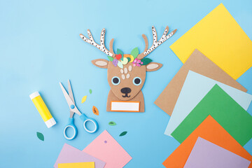 Fototapeta premium Reindeer head craft with floral crown, scissors, glue, and colored paper on light blue background for kids workshops, DIY product packaging, craft kit stores, educational supply business use,