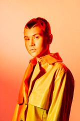 young woman with short hair wearing a yellow jacket and posing with a serious expression against an...