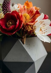 A vibrant bouquet of colorful flowers, including red, orange, and white blooms, arranged in a modern, geometric concrete vase on a dark surface.