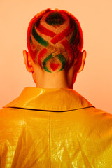 Woman with colorful geometric shaved haircut wearing a textured yellow jacket, standing against a...