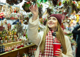 Young woman walks with disposable cup of coffee and view hanging toys near window of New Year...
