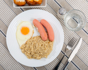 Two sausages with a fried egg and oatmeal on a white plate. Breakfast option with eggs and porridge...