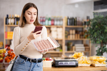 Young woman buyer scanning qr code of raw sausages with phone in grocery store