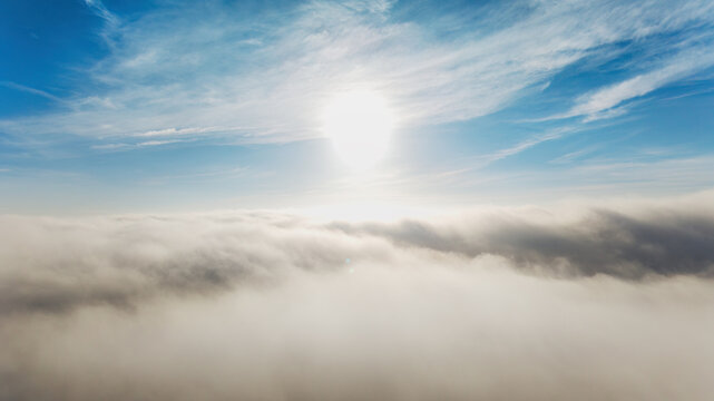Bright sunlight pierces through fluffy clouds, illuminating a serene blue sky above a sea of misty clouds below.