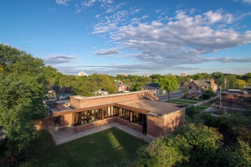 A delightful brick building nestled in lush greenery, set within a tranquil neighborhood. The scene is graced by a bright blue sky and fluffy white clouds.