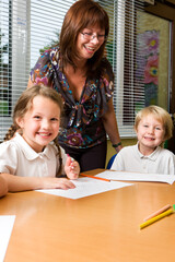 A smiling female teacher stands behind two happy children in a school classroom. The image captures a candidly positive and supportive relationship between a teacher and her pupils. From a series.