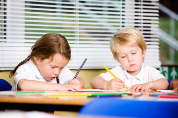 Two school children colouring in class. A young girl and boy sit at their desk, intently focused on drawing or colouring with coloured pencils on a piece of paper. From a series of images.