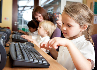 Children working on computers in a classroom. A young primary girl is focused on typing on a computer keyboard during a lesson with her teacher and other classmates.