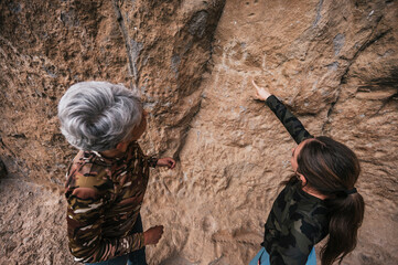 Mother and daughter dressed in hiking clothes, walking along a cliff observing cave paintings.	
