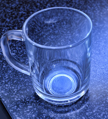 A transparent glass mug with a massive handle stands on a dark tabletop with a textured pattern. 