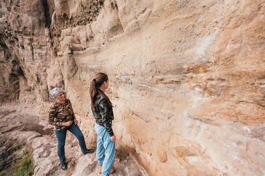 Mother and daughter dressed in hiking clothes, walking along a cliff observing cave paintings.	
