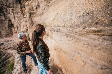 Naklejka premium Mother and daughter dressed in hiking clothes, walking along a cliff observing cave paintings. 