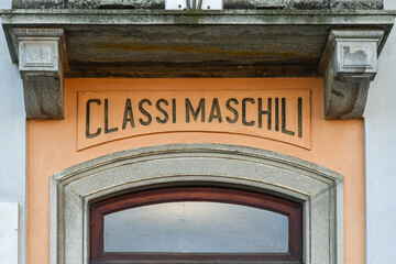Close-up of an old, obsolete sign that says: "Classi Maschili" ("Male Classes") on the facade of an elementary school, Italy