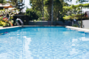 Naklejka premium A beautifully blurred swimming pool with blue tiles and sparkling water, surrounded by lush greenery, ideal for a serene summer escape or luxurious travel.