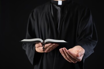 Priest with Holy Bible and rosary preaching on black background, closeup