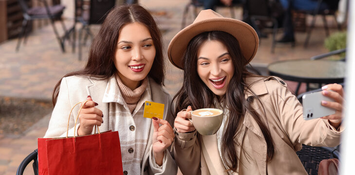 Female friends with shopping bag and coffee taking selfie at table in street cafe
