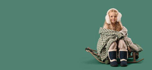 Cute little girl in winter clothes and with warm plaid sitting on sled against green background...