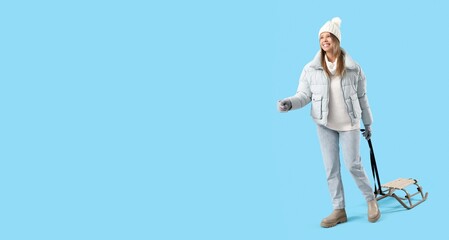 Happy young woman in winter clothes and with sled on blue background with space for text