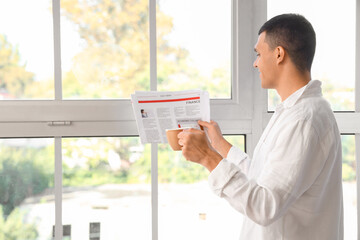Handsome young man holding cup of coffee reading newspaper near window at home