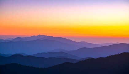 A vibrant sunset casts a golden glow over layers of hazy blue mountains, creating a serene vista.