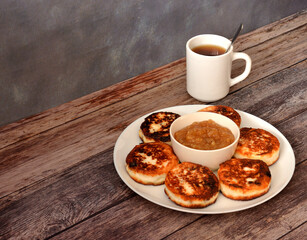 A large plate of fresh cheesecakes with raisins, a cup of apple jam and hot tea on a wooden table.
