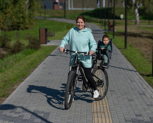 Caucasian woman riding bicycle with toddler son sitting behind her in child seat in park. 