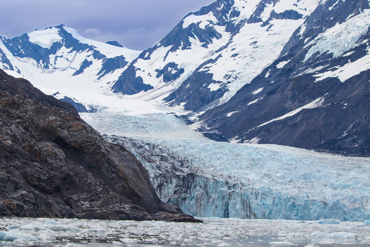 A stunning view of Alaska's Surprise Glacier. The massive blue and white ice field flows between two rugged, snow-capped mountains and calves into the water below, filled with floating ice.