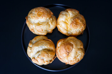Top view of a group of freshly baked Kue Sus (Choux Pastries or Cream Puffs), with a beautiful golden-brown exterior, arranged on a dark plate.