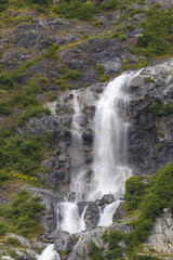 A majestic waterfall tumbles down a sheer, jagged rock face covered in green foliage and moss. The powerful white water contrasts with the dark stone, creating a stunning Alaskan scene.