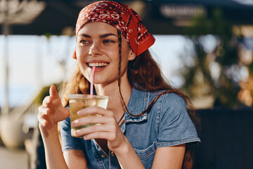 Smiling woman wearing a red bandana and denim shirt enjoying a refreshing drink outdoors with warm...