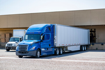 Big rig blue semi truck with dry van semi trailer and middle duty white day cab semi truck with box trailer standing side by side in warehouse dock gates loading cargo for next freights deliveries
