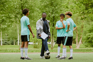 Black man coaching three teenage boys of diverse ethnicity standing on grass field, holding...