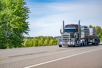 Classic black big rig semi truck with aluminum protective grille guard and chrome accessories transporting fastened lumber wood on flat bed semi trailer driving on the flat highway road