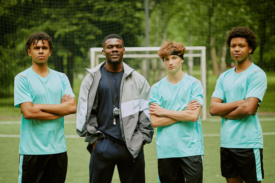 Black young adult man standing with arms relaxed holding whistle surrounded by three multiethnic teenage boys crossing arms on outdoor soccer field - Powered by Adobe