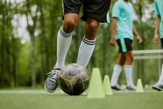 Teenager boys practicing soccer dribbling skills on outdoor field, focusing on controlling ball around training cones, legs and lower bodies visible, teammates standing in background